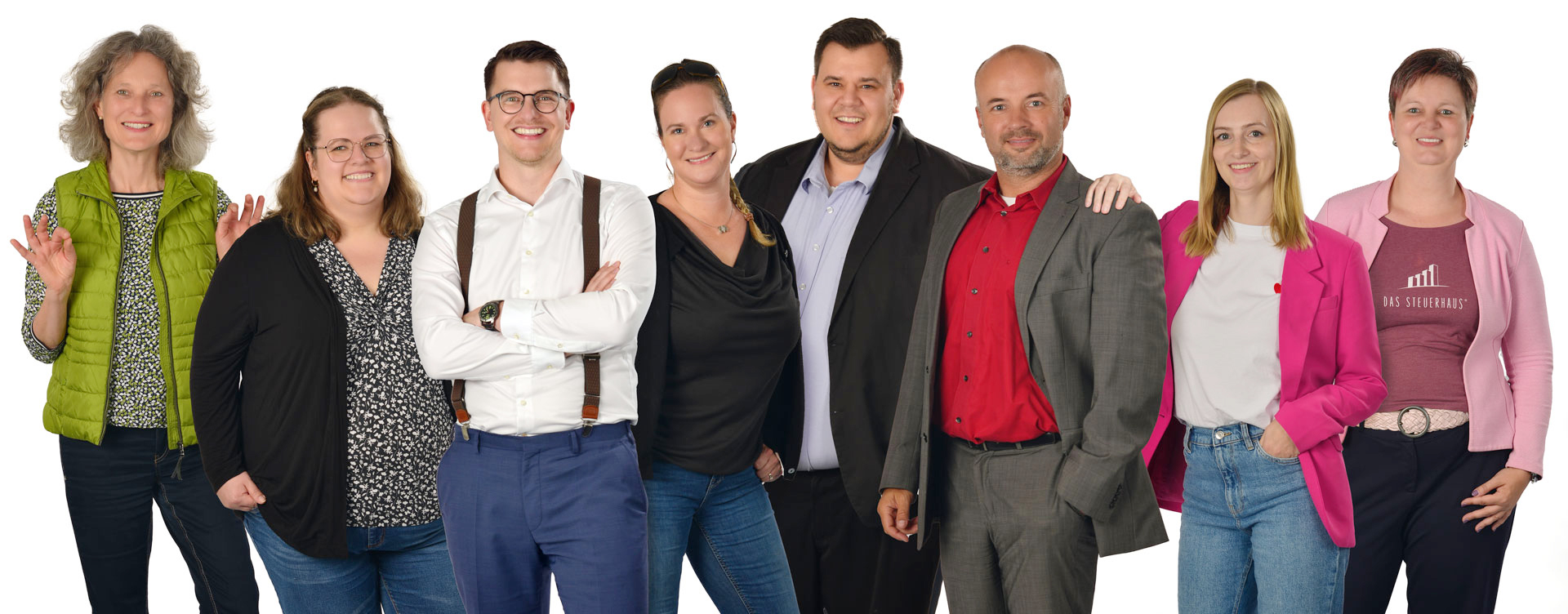 Das Bild zeigt ein Gruppenfoto mit acht Personen, die nebeneinander vor einem weißen Hintergrund aufgereiht stehen. Die Gruppe besteht aus Männern und Frauen in unterschiedlicher Business-Kleidung. Links steht eine Frau mit lockigem Haar in einer grünen Steppjacke, während andere Personen Anzüge, Hemden und Blazer in verschiedenen Farben tragen. Die Personen in der Mitte tragen formellere Kleidung, darunter ein Mann mit weißem Hemd und Hosenträgern sowie mehrere Personen in dunklen Sakkos. Rechts stehen zwei Frauen in auffälligen rosa/pinken Blazern. Alle lächeln in die Kamera und wirken wie ein professionelles Team oder eine Arbeitsgruppe.