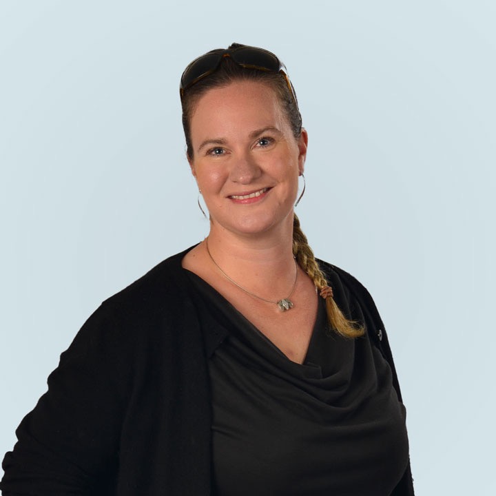 Eine Frau mit blonden Zöpfen und Sonnenbrille auf dem Kopf lächelt freundlich in die Kamera, trägt ein schwarzes Top und eine Kette auf hellem Hintergrund.[5]