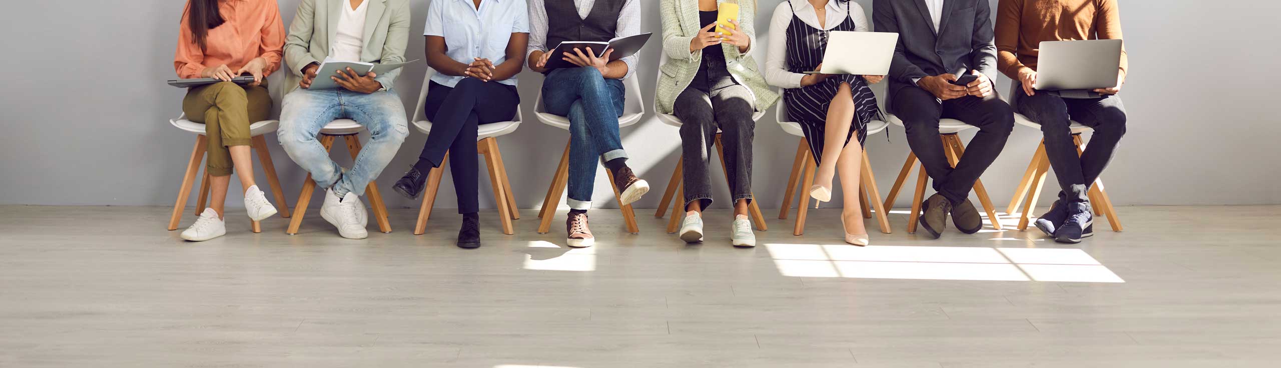 Das Bild zeigt eine Reihe von Personen, die nebeneinander auf weißen Stühlen mit Holzbeinen sitzen. Sie sind unterschiedlich gekleidet - einige in Businesskleidung, andere in legereren Outfits. Die meisten beschäftigen sich mit elektronischen Geräten wie Laptops, Tablets oder Smartphones, während eine Person in einem Notizbuch liest. Die Szene spielt vor einer schlichten grauen Wand auf einem hellen Holzboden, wobei Sonnenlicht durch ein nicht sichtbares Fenster auf den Boden fällt. Die Situation erinnert an Menschen in einem Wartezimmer oder Wartebereich, die sich während des Wartens mit verschiedenen Aktivitäten beschäftigen.