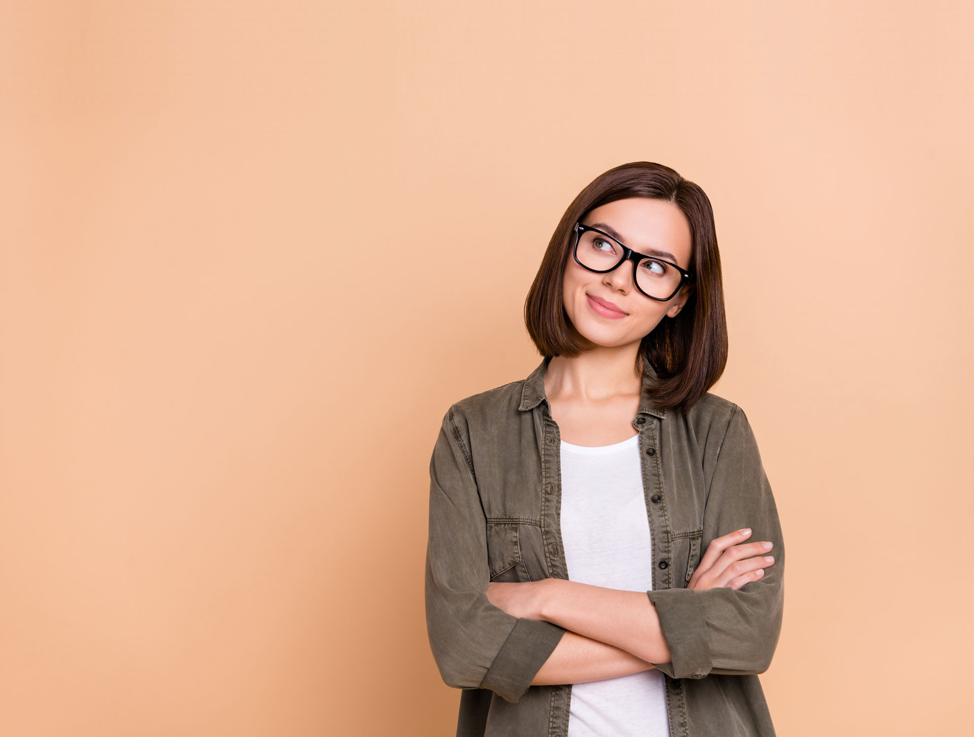 Das Bild zeigt eine Person mit schulterlangem braunem Haar, die eine schwarze, markante Brille trägt. Sie blickt nachdenklich zur Seite und lächelt leicht. Die Person trägt ein olivgrünes Hemd über einem weißen Oberteil und hat die Arme vor der Brust verschränkt. Der Hintergrund ist in einem warmen Pfirsichton gehalten, der einen schönen Kontrast zur Kleidung bildet. Die Gesamtkomposition wirkt freundlich und professionell.